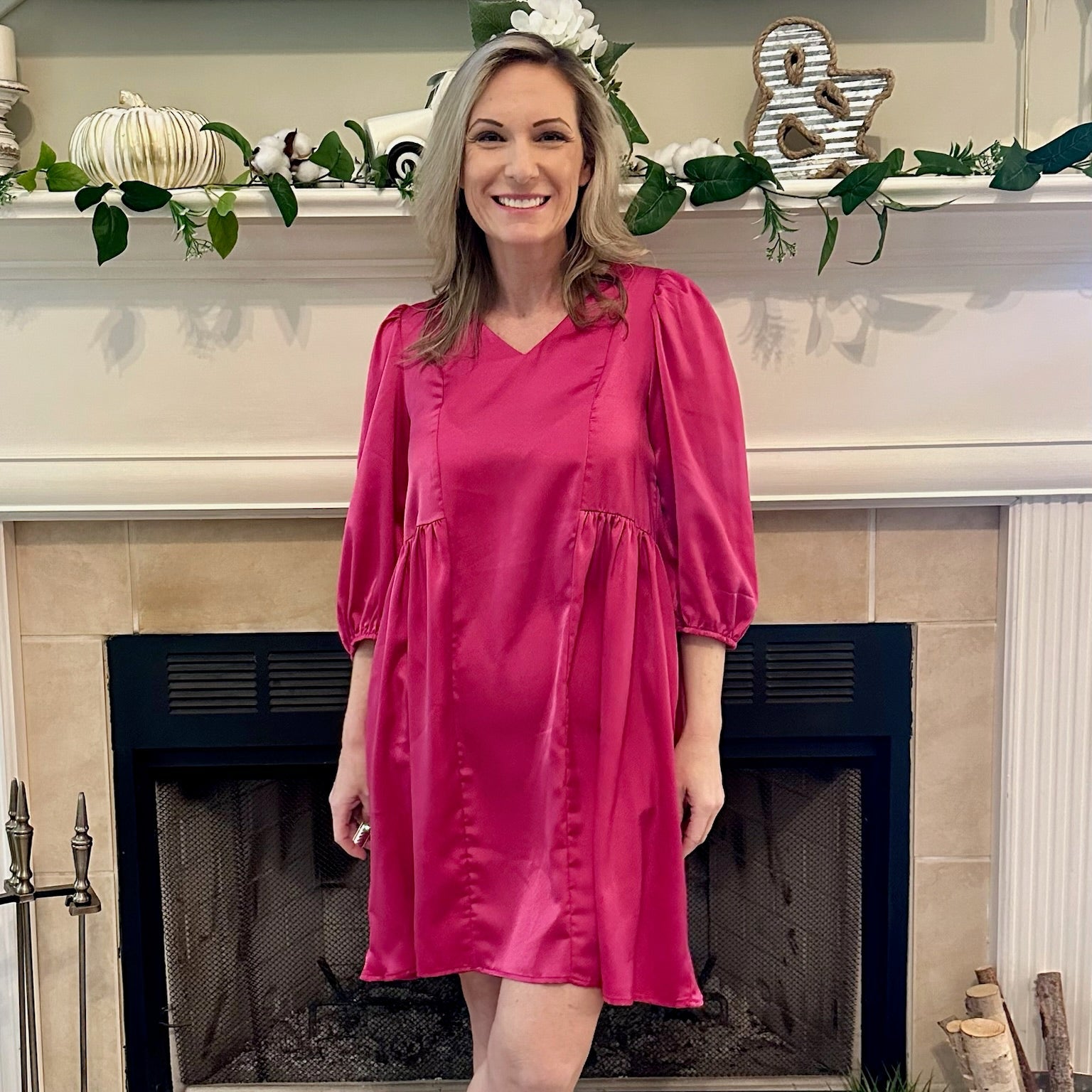 Woman in a pink dress standing in front of a fireplace with decorative items.