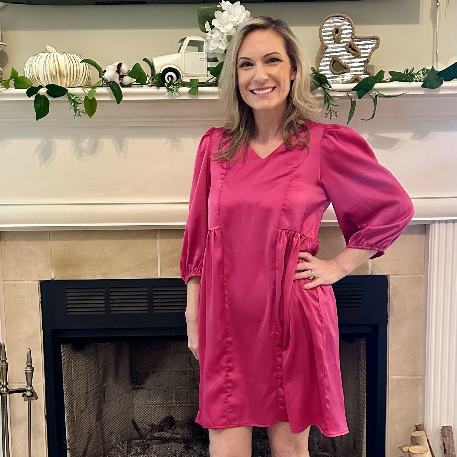 Woman in a pink dress standing in front of a fireplace with decorative items.