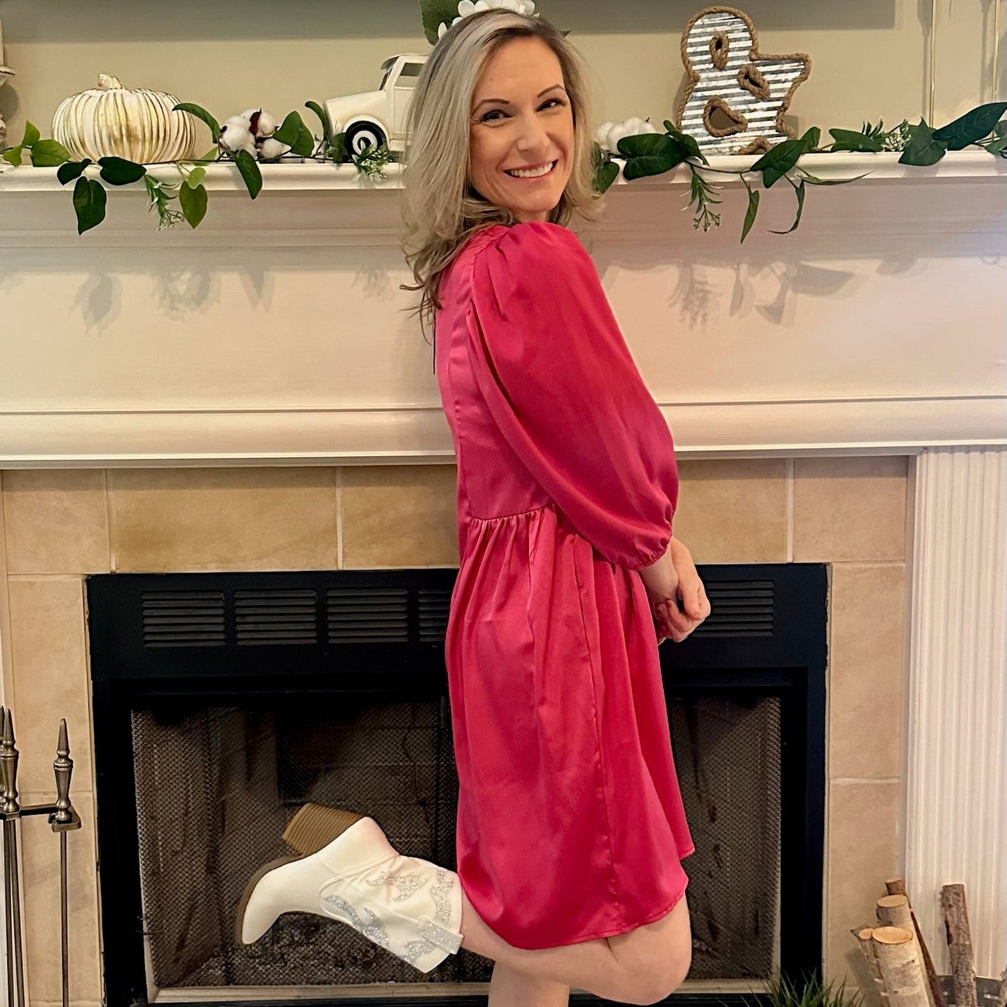 Woman in a pink dress and white boots standing in front of a fireplace with holiday decorations.