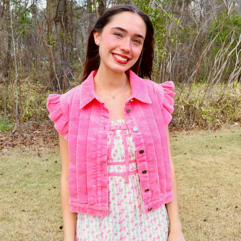 A young woman smiling outdoors in a grassy wooded area, wearing a bright pink corduroy ruffle-sleeve button-up vest over a white and pink floral dress.
