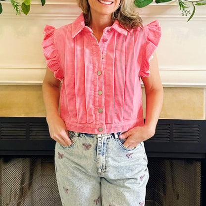 A woman wearing a pink corduroy button-up shirt with ruffled sleeves and light-wash denim jeans, standing in front of a fireplace.
