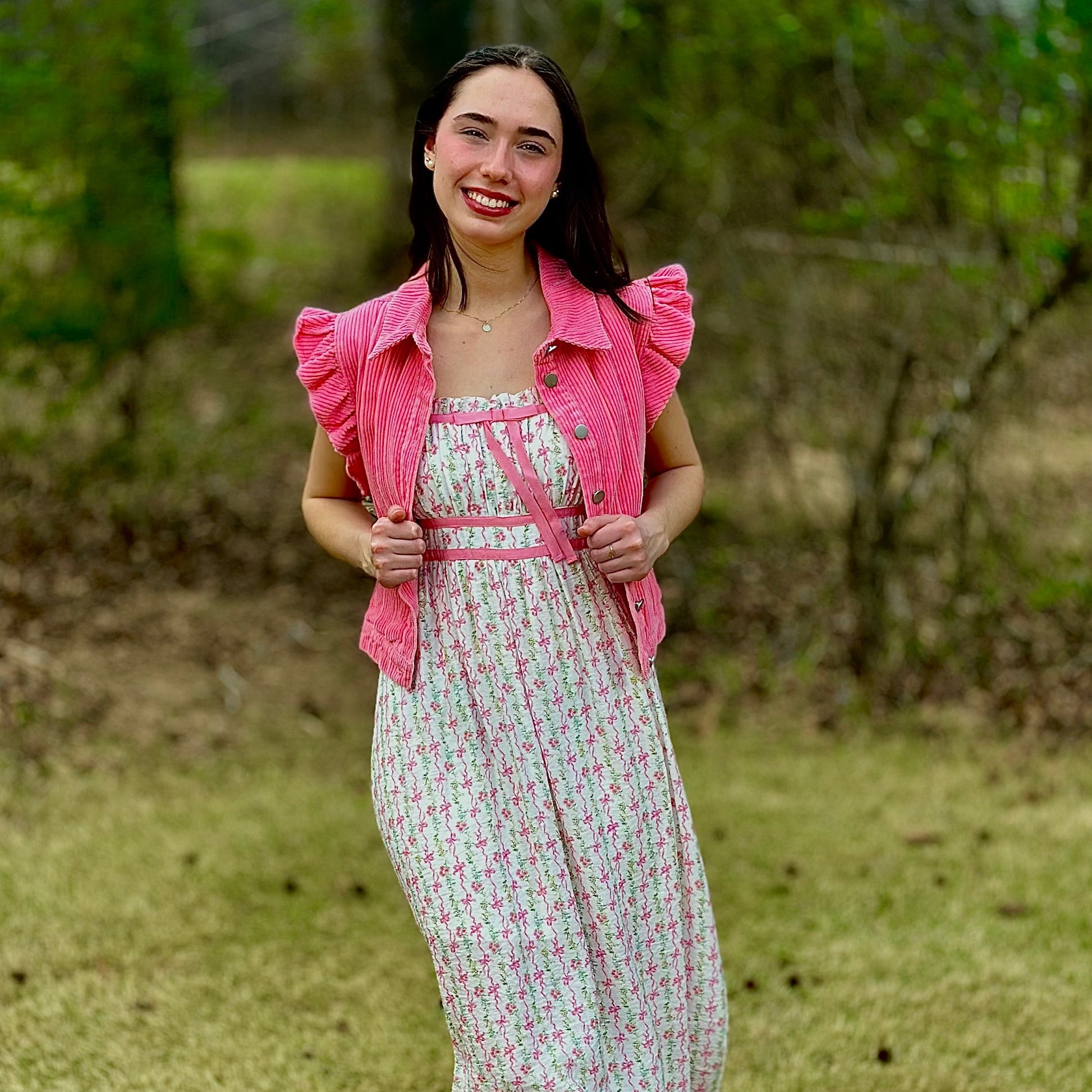 A woman stands outdoors on grass wearing a white midi dress with a pink floral print, a matching pink ruffled jacket, and white flats.