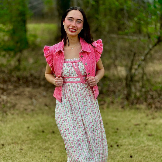 A woman stands outdoors on grass wearing a white midi dress with a pink floral print, a matching pink ruffled jacket, and white flats.