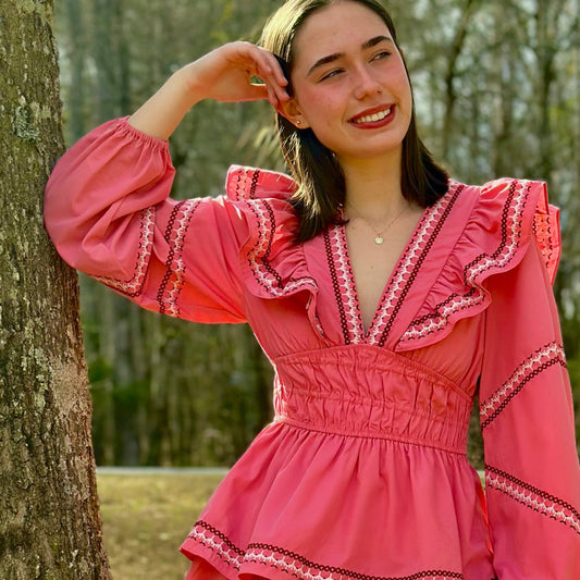 A young woman wearing a pink tiered mini dress with ruffled details and long puff sleeves, posing outdoors next to a large tree trunk in a wooded area.
