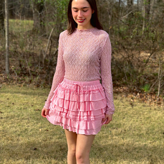 A woman wearing a pink long-sleeve knit top and a pink ruffled tiered mini skirt, standing outdoors on a grassy lawn with trees in the background.