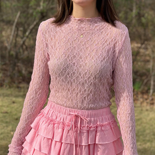 A young woman wearing a light pink long-sleeve lace top and a matching pink tiered ruffle mini skirt, standing outdoors in a grassy field with trees in the background.