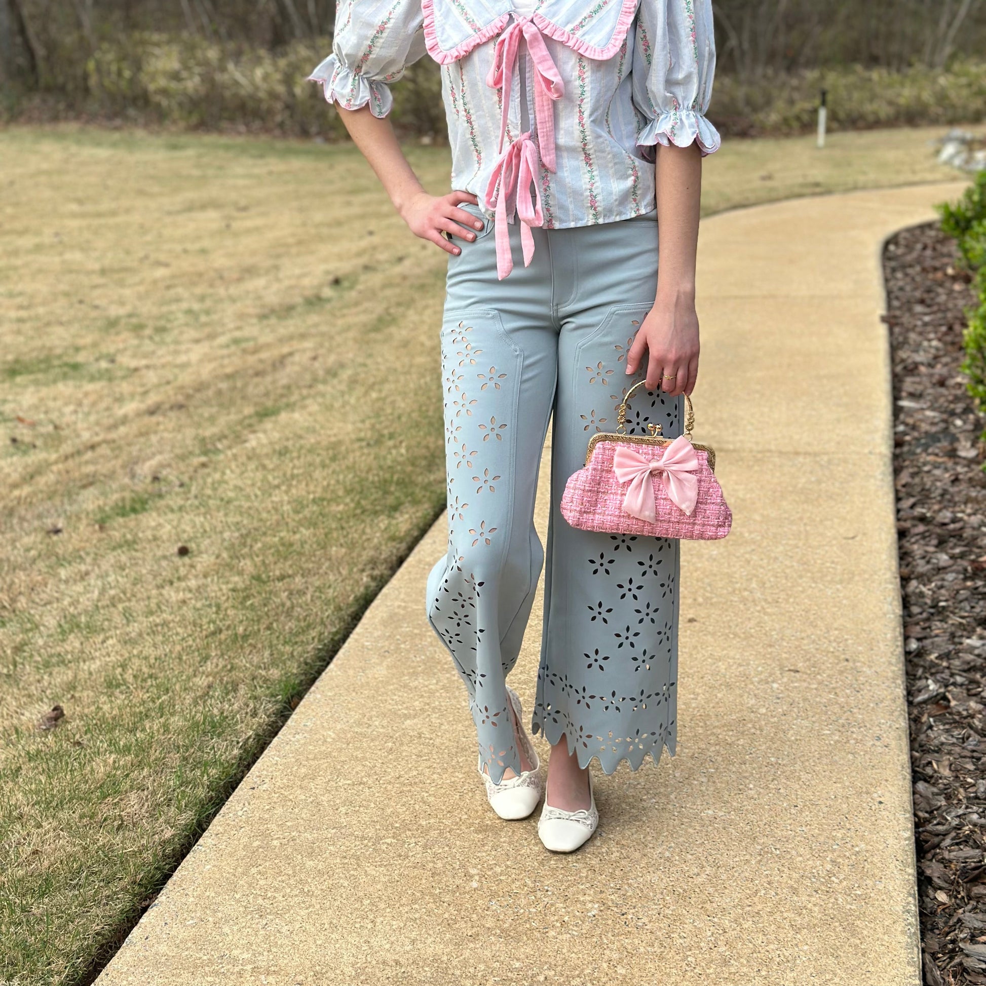 A woman stands on a paved path outdoors wearing a white ruffled blouse with pink trim and a matching pink bow tie, paired with light gray eyelet pants, white flats, and holding a pink quilted handbag.