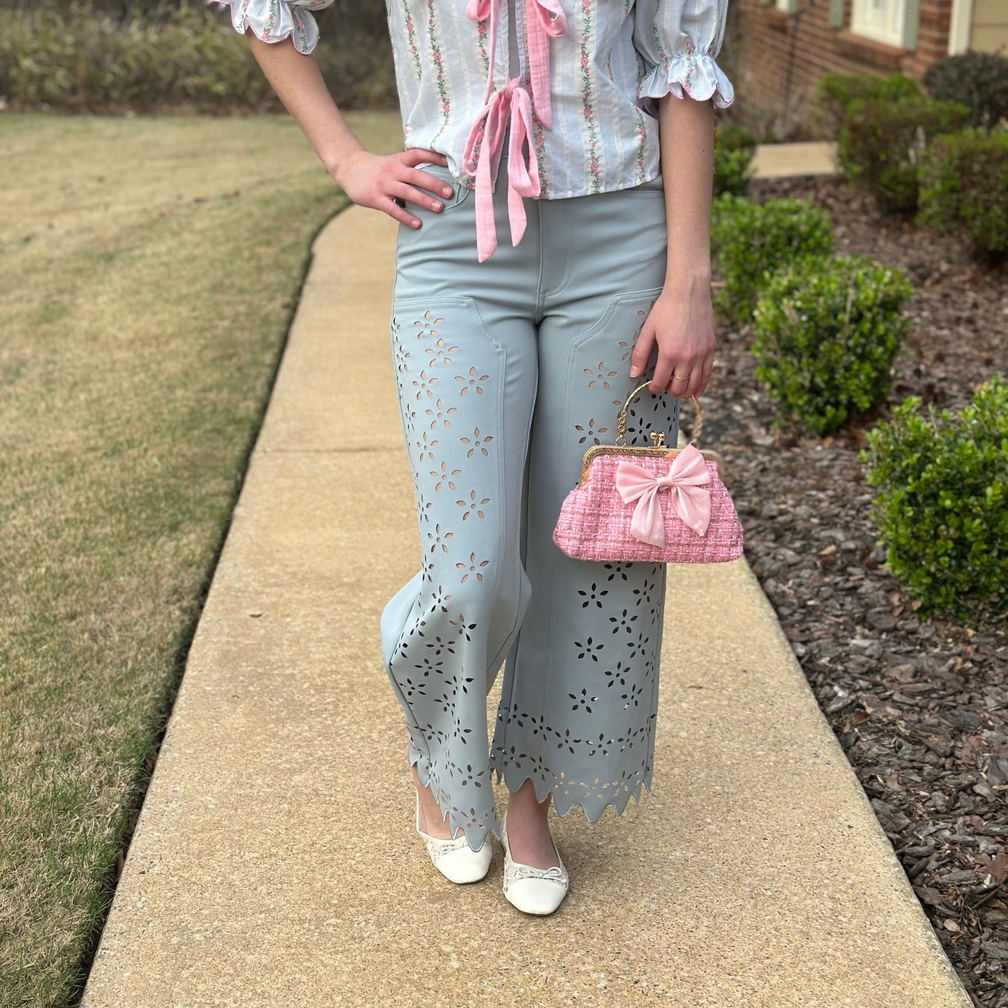A person standing on a concrete walkway outdoors wearing a light blue and white striped blouse with pink ruffle trim and tie details, matching light blue eyelet pants, white shoes, and holding a pink quilted handbag.