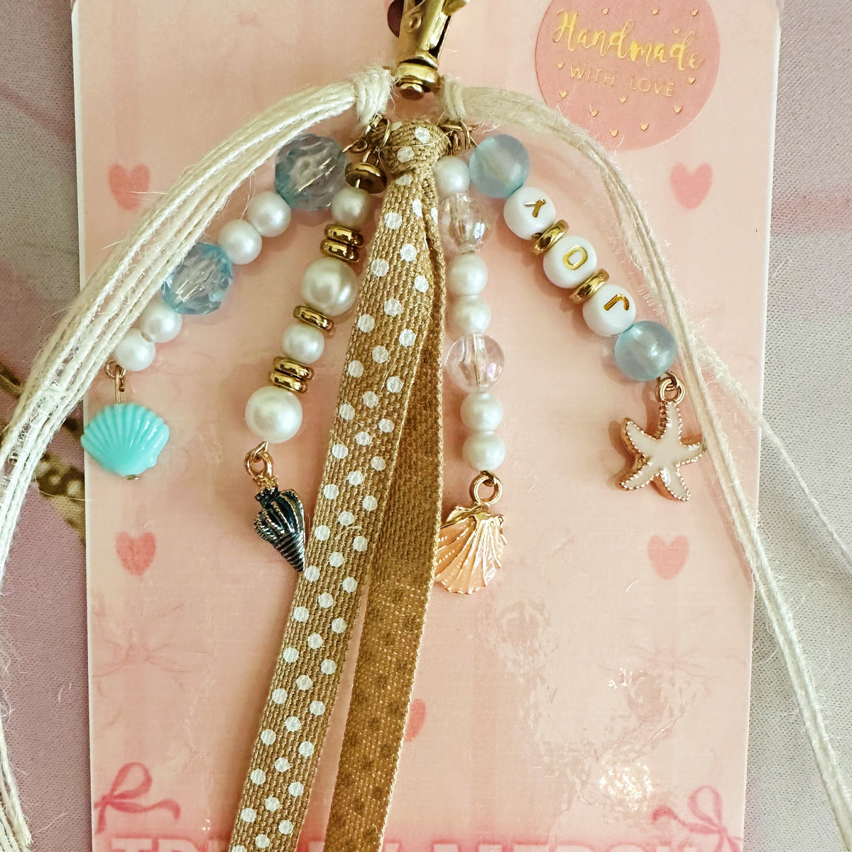 A handmade beaded keychain with a gold clasp, featuring light blue and white beads, letter beads spelling 'JOY', a pink starfish charm, and small shell charms, displayed on a wooden surface.