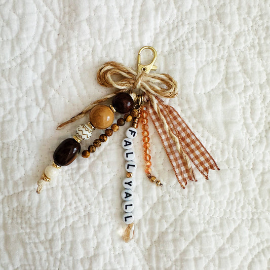 A handmade autumn-themed beaded keychain charm with a gold clasp, wrapped in twine bow, featuring brown and beige beads, a white bead spelling 'FALL YA'LL and a brown gingham ribbon tassel, displayed on a white quilted blanket.