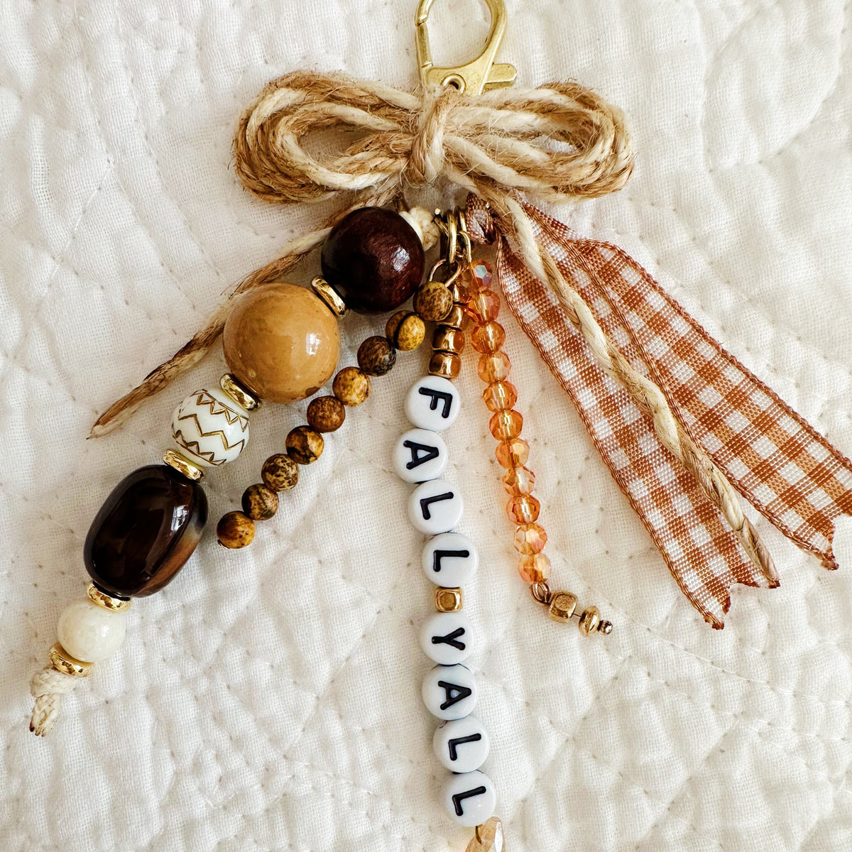 A handmade autumn-themed keychain or bag charm with a rustic twine bow, wooden beads, amber beads, a white beaded 'FALL/YALL' charm, and an orange-and-white gingham ribbon, displayed on a white quilted blanket.