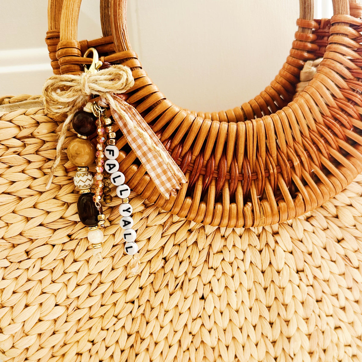 A close-up of a woven straw tote bag with a curved wicker handle, decorated with a beaded charm that reads 'FALL YA'LL' and a small gingham ribbon bow.