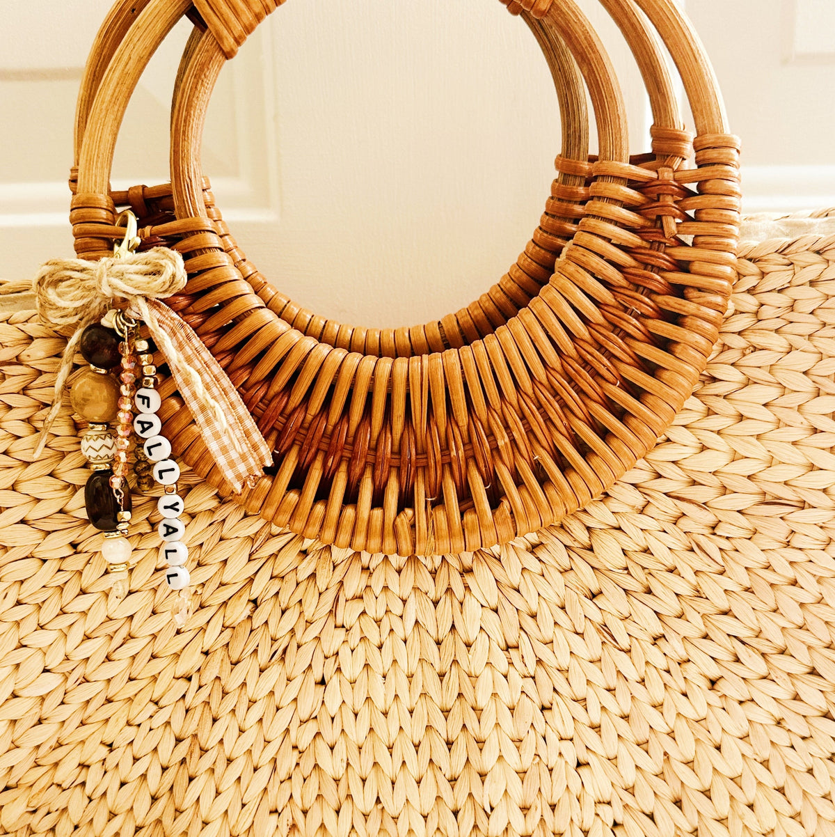 A natural straw tote bag with a wide, curved double-ring handle made of woven rattan, featuring a decorative charm with beads and a tassel that reads 'FALL YA'LL. 