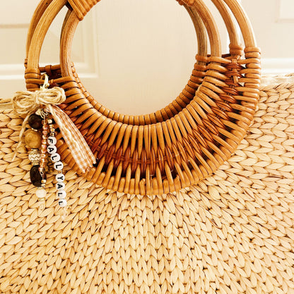A natural straw tote bag with a wide, curved double-ring handle made of woven rattan, featuring a decorative charm with beads and a tassel that reads 'FALL YA'LL. 