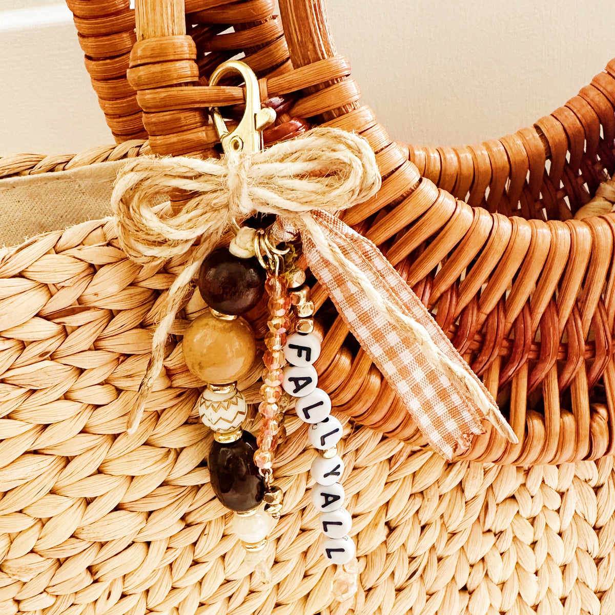 A close-up of a woven straw tote bag with a decorative keychain charm tied to the handle. The charm includes a gold clasp, a jute ribbon bow, wooden beads, a small gingham ribbon accent, and white letter beads spelling 'FALL YA'LL. 