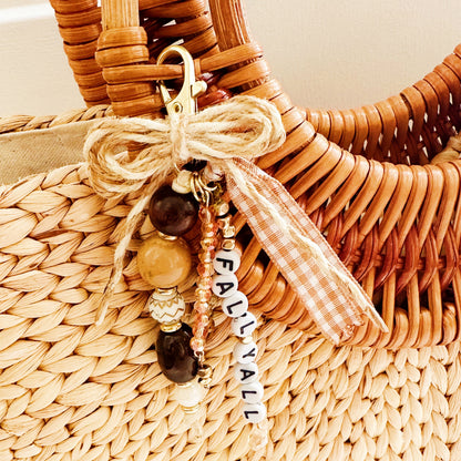 A close-up of a woven straw tote bag with a decorative keychain charm tied to the handle. The charm includes a gold clasp, a jute ribbon bow, wooden beads, a small gingham ribbon accent, and white letter beads spelling 'FALL YA'LL. 