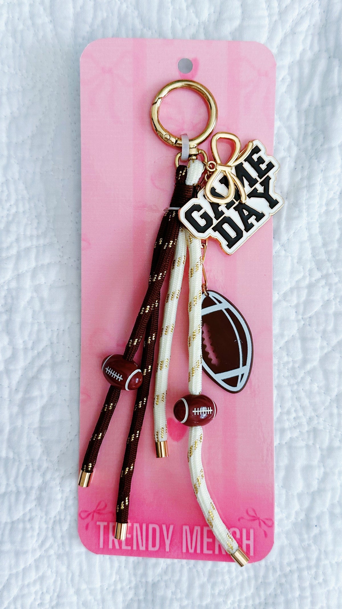 A football themed keychain set on a pink display card. The keychain features a gold-tone split ring, a 'GAME DAY' charm, a football-shaped charm, and three dangling rope tassels in brown and cream with gold speckles and small brown footballs. 