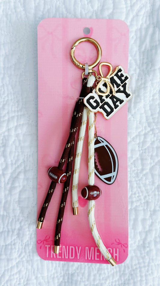 A football themed keychain set on a pink display card. The keychain features a gold-tone split ring, a 'GAME DAY' charm, a football-shaped charm, and three dangling rope tassels in brown and cream with gold speckles and small brown footballs. 