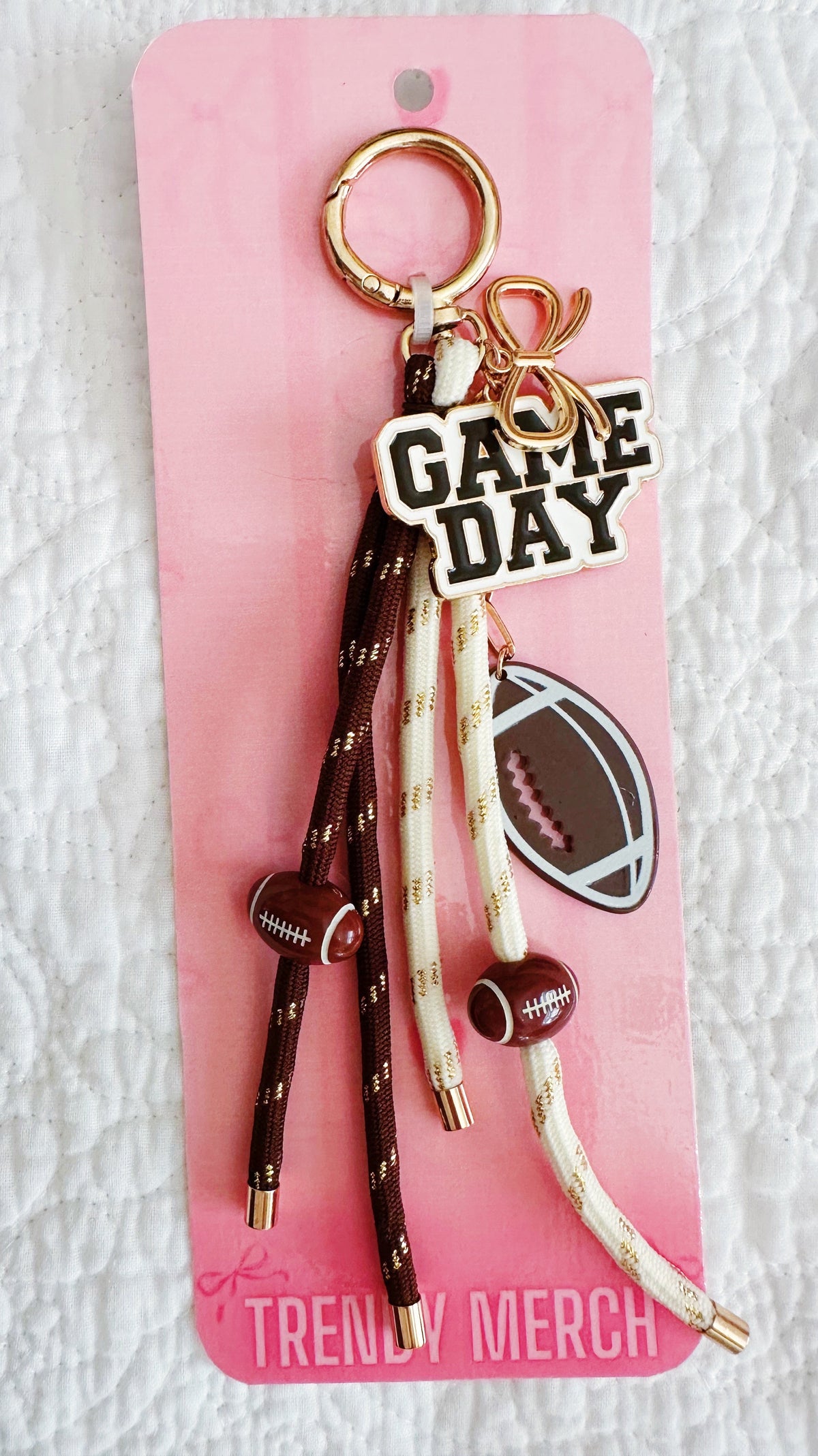 A 'Game Day' themed keychain with football-themed charms, including a football-shaped charm, a football graphic, and brown and cream rope tassels, attached to a pink card labeled 'Trendy Merch'.