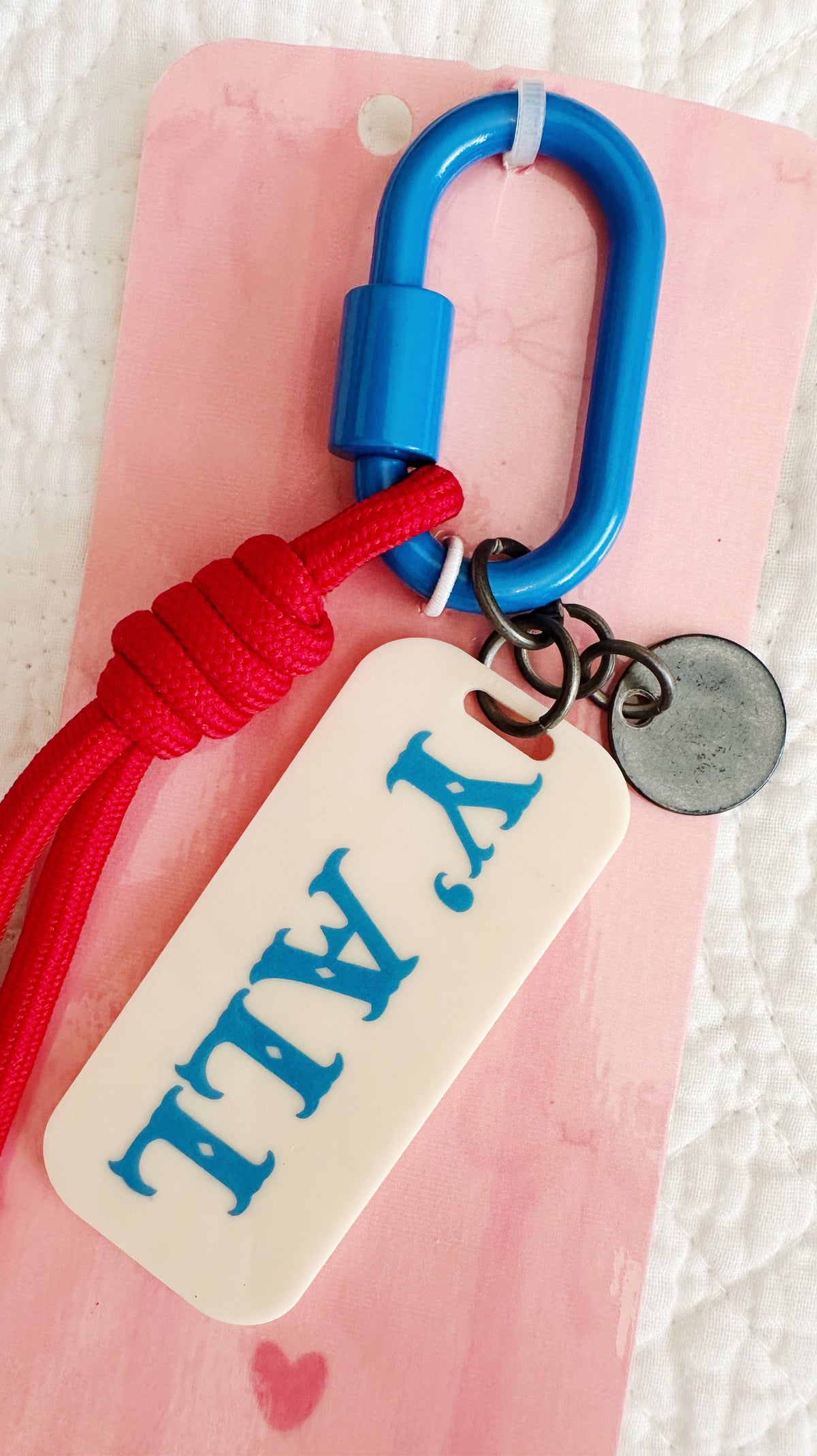 A close-up of a keychain or bag charm with a bright blue carabiner, red braided cord, and an off-white rectangular tag printed with 'Y'ALL' in blue. The item is displayed on a pink backing card over a white textured fabric background.