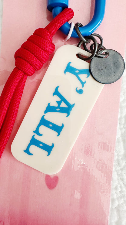 A close-up of a keychain or bag charm featuring a red braided cord, a blue carabiner, and a cream-colored rectangular tag with the word 'Y'ALL' in blue. A small round metal tag is attached.