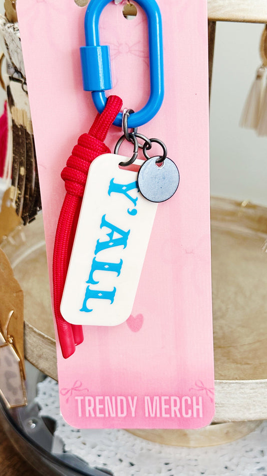A close-up of a keychain or bag charm attached to a pink retail card. The product has a bright blue carabiner clip, a red knotted cord, a white rectangular tag with blue text reading 'Y'ALL', and a small round silver metal tag.