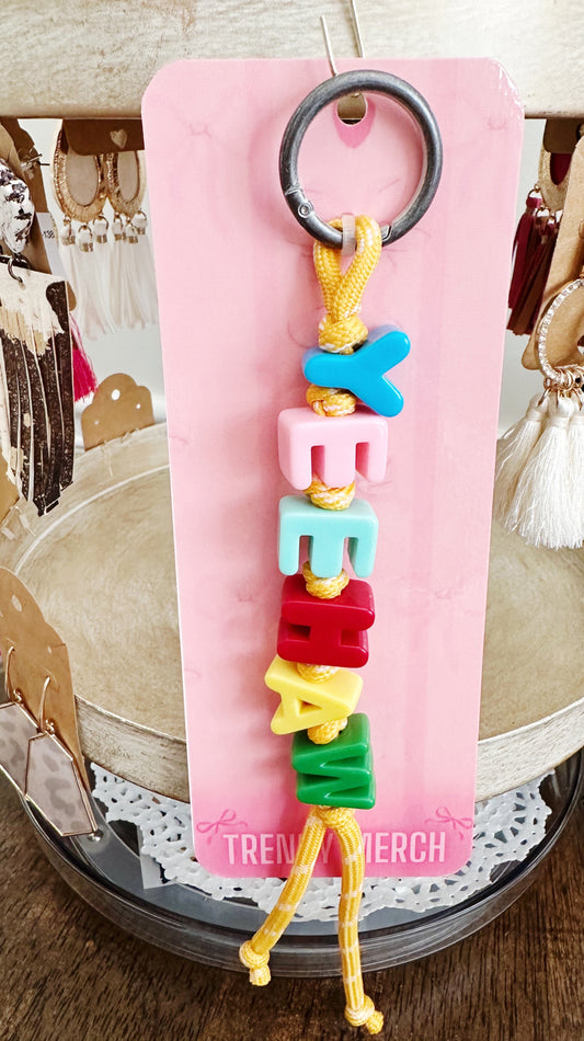 A colorful beaded keychain with the word 'MAMMA' spelled out in pastel pink, blue, light blue, red, yellow, and green beads, hanging from a black metal ring on a pink display card.