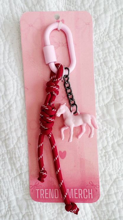 A pink and red keychain or bag charm with a pink horse charm, red braided cord, and black chain, attached to a pink retail card labeled 'TREND MERCH'.