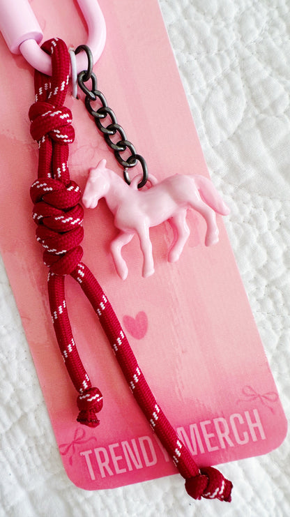 A pink and red keychain or bag charm featuring a knotted red cord, a small pink unicorn charm, and a black metal chain, attached to a pink retail card labeled TREND MERCH.