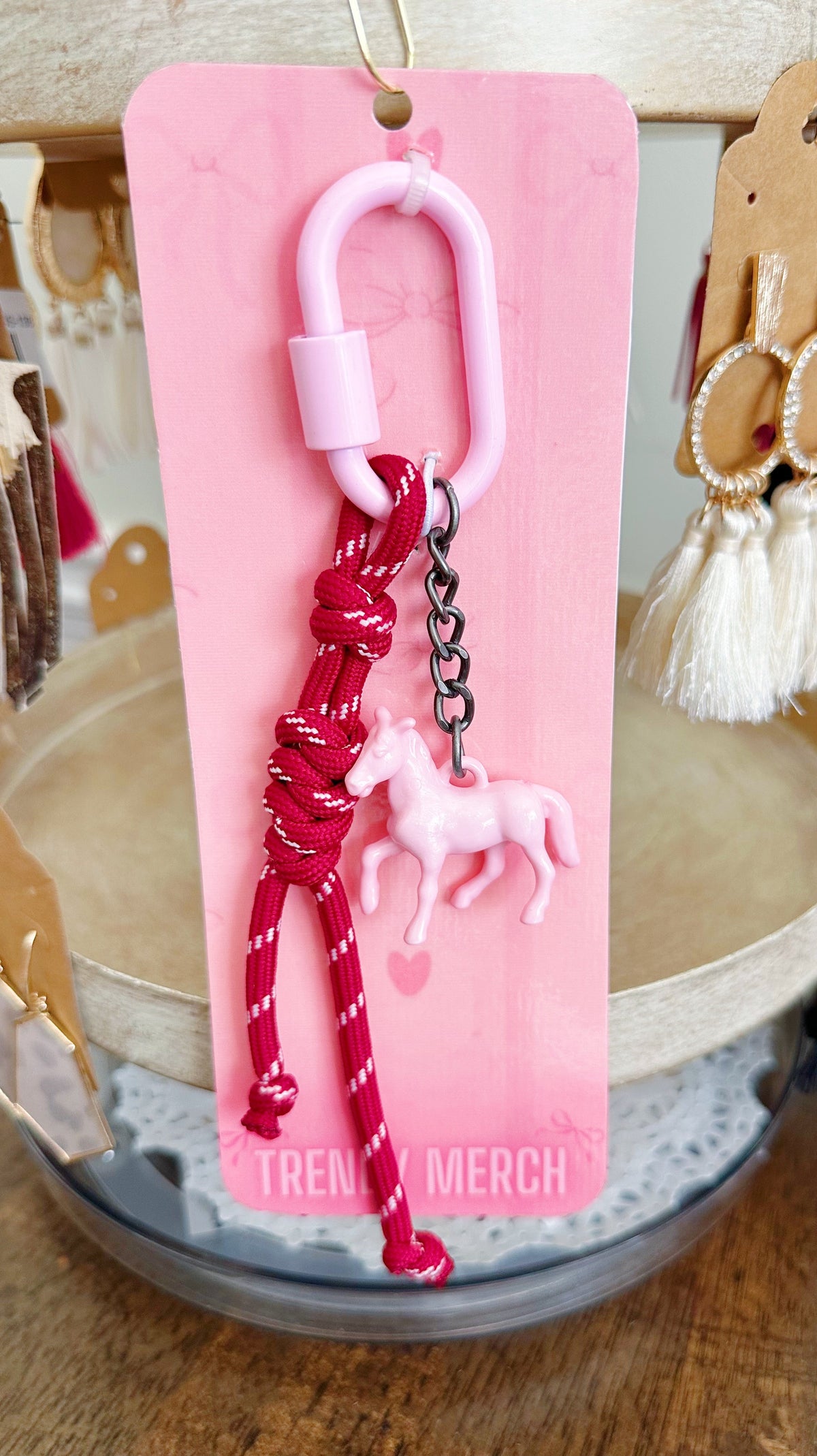 A pink and red keychain accessory with a small pink horse charm, hanging from a pink display card on a wooden surface.