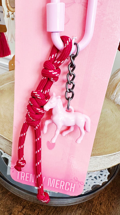 A pink and red keychain accessory attached to a pink display card. The keychain features a red braided cord with white accents, a black metal chain, and a small white unicorn charm.