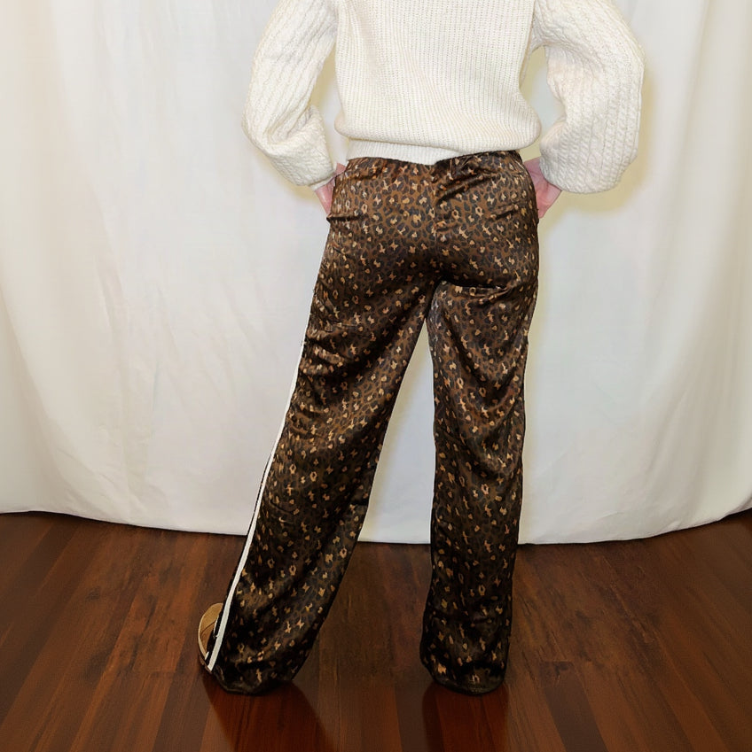 A person shown from the back wearing brown patterned wide-leg pants with a white side stripe and a cream ribbed sweater, standing against a white draped backdrop on a wooden floor.