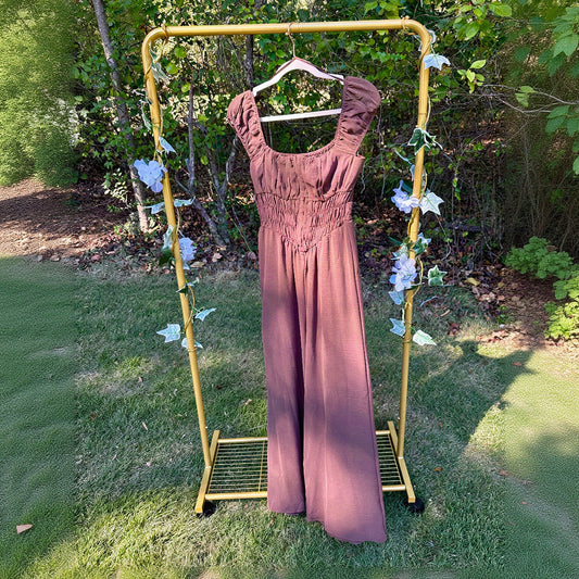 A brown jumpsuit with puff sleeves and a smocked bodice hangs on a yellow rolling clothing rack outdoors on a grassy lawn with trees in the background.