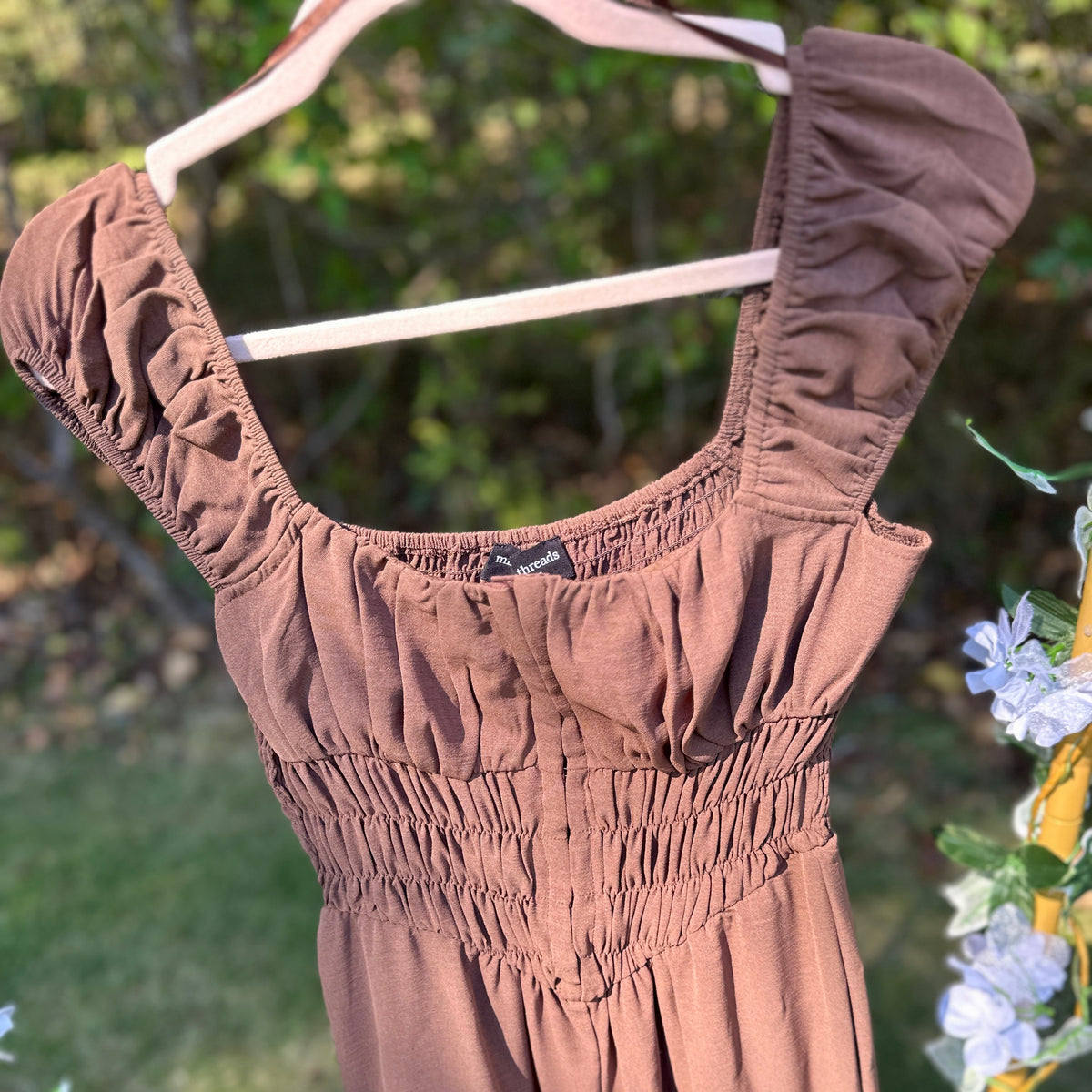 A brown smocked jumpsuit with ruffled puff sleeves, hanging on a white hanger against a blurred outdoor garden background.