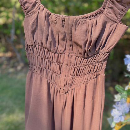 A brown smocked jumpsuit with ruffled shoulder details, shown hanging outdoors against a blurred green garden background.