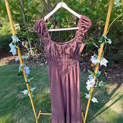 A brown jumpsuit with a smocked bodice and ruffled cap sleeves, hanging on a white hanger from a yellow garden arch decorated with artificial flowers, set against a green garden background.