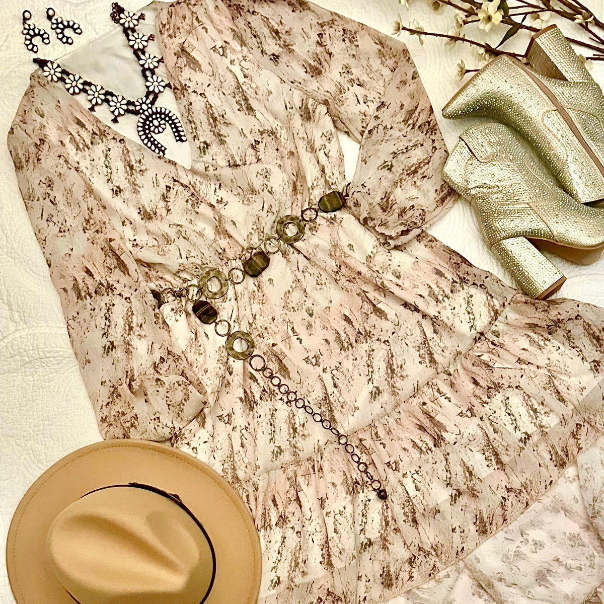 A beige and brown patterned long-sleeve dress styled in a flat lay with a tan hat, a silver statement necklace, a gold belt, and metallic gold high-heeled boots on a white textured fabric background.