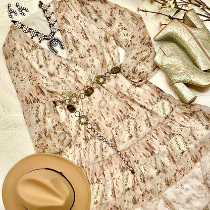 A beige and brown patterned long-sleeve dress styled in a flat lay with a tan hat, a silver statement necklace, a gold belt, and metallic gold high-heeled boots on a white textured fabric background.
