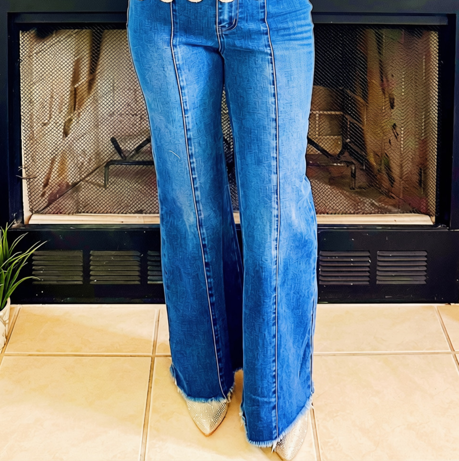 A person wearing blue flare jeans with frayed hems and pointed-toe metallic boots, standing in front of a fireplace with tiled flooring.