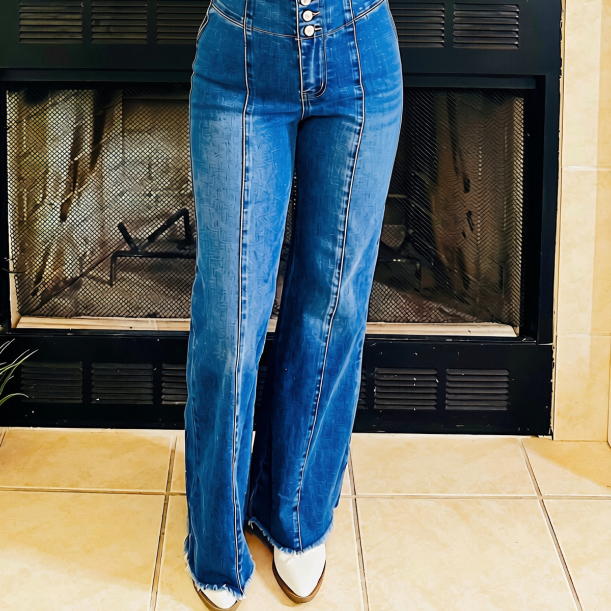 A person wearing high-waisted, wide-leg blue denim jeans with a button fly and frayed hems, paired with white pointed-toe boots, standing in front of a fireplace.