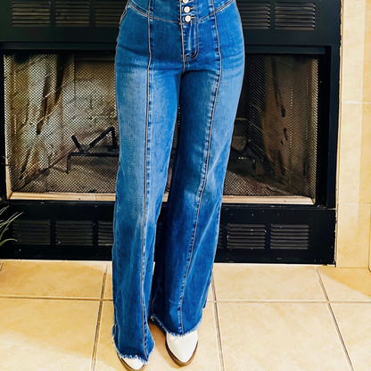 A person wearing high-waisted, wide-leg blue denim jeans with a button fly and frayed hems, paired with white pointed-toe boots, standing in front of a fireplace.