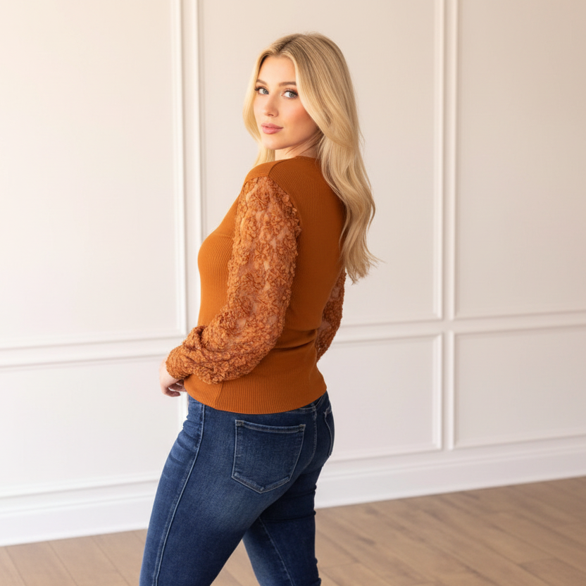 Woman wearing a brown sweater and blue jeans standing against a white paneled wall.